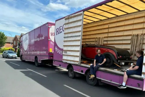 Regal Moving & Storage purple removal truck with side door open and a vehicle loaded, crew sitting on the side